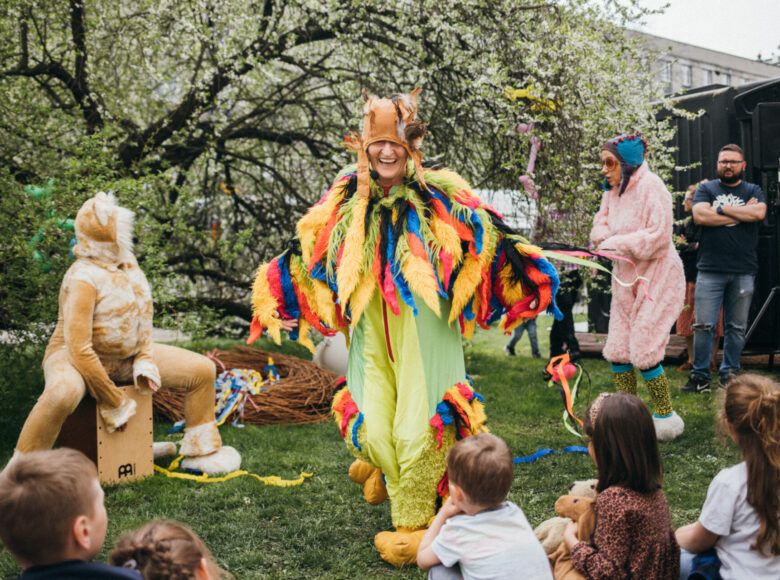 Zdjęcie. Aktorzy znajdują się w parku. Są przebrani za kolorowe ptaki. Grają spektakl. Dzieci siedzą na trawie i oglądają sztukę.