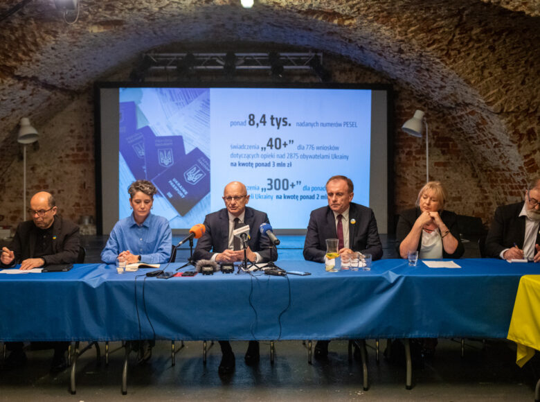 Zdjęcie. Za stołem okrytym niebieskim obrusem siedzi grupa osób. Na stole lezą radiowe mikrofony, karafka z woda i szklanki. W tle wyświetla się prezentacja.