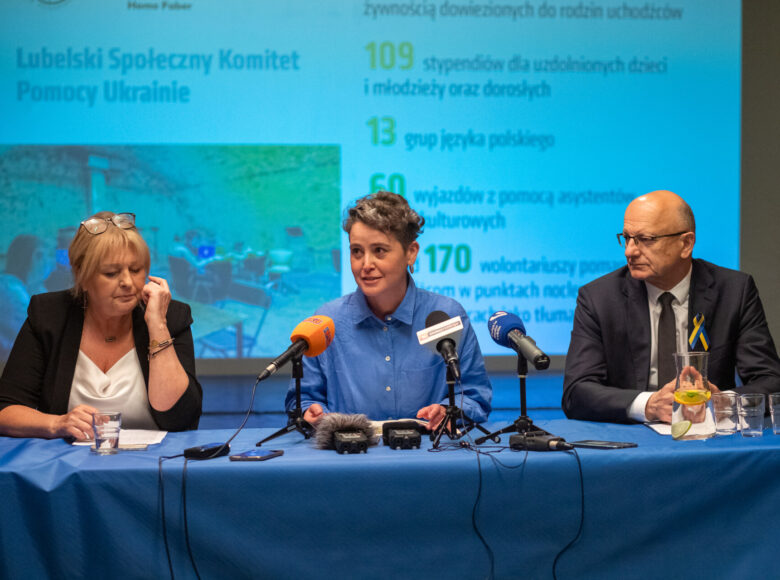 Zdjęcie. Za stołem okrytym niebieskim obrusem siedzą dwie kobiety i mężczyzna. Na stole lezą radiowe mikrofony, karafka z woda i szklanki. W tle wyświetla się prezentacja.