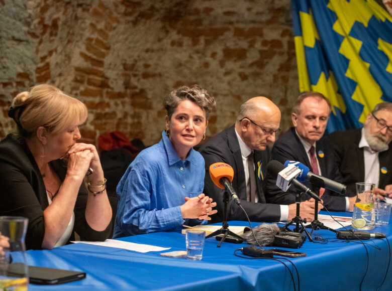 Zdjęcie. Za stołem okrytym niebieskim obrusem siedzi grupa osób. Na stole lezą radiowe mikrofony, karafka z woda i szklanki. Na ścianie wisi chusta z niebiesko-żółtym wzorem.