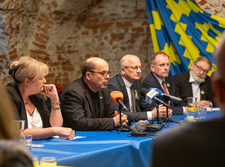 Zdjęcie. Za stołem okrytym niebieskim obrusem siedzi grupa osób. Na stole lezą radiowe mikrofony, karafka z woda i szklanki. Na ścianie wisi chusta z niebiesko-żółtym wzorem.