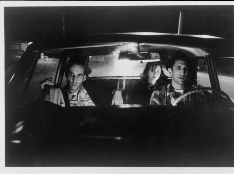 Czarno-białe zdjęcie. Trzy osoby siedzą w samochodzie – dwóch mężczyzn z przodu, kobieta za nimi.