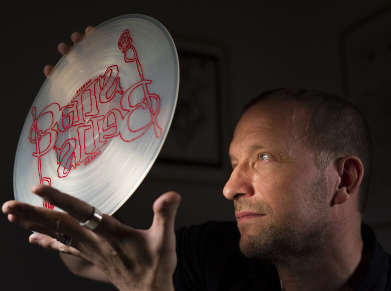 Zdjęcie w orientacji poziomej, kolorowe. Na czarnym tle, z prawej strony widoczna jest głowa białego mężczyzny, z krótkimi jasnymi włosami i lekkim zarostem, zwrócone nieco w lewo. Mężczyzna trzyma w wyciągniętych i lekko uniesionych dłoniach płytę winylową, wykonaną z jasnego transperentnego materiału. Na płycie znajduje się podwojony czerwony napis „Bells” – ozdobnymi literami oraz stereo – mniejszym krojem pisma, wersalikami.