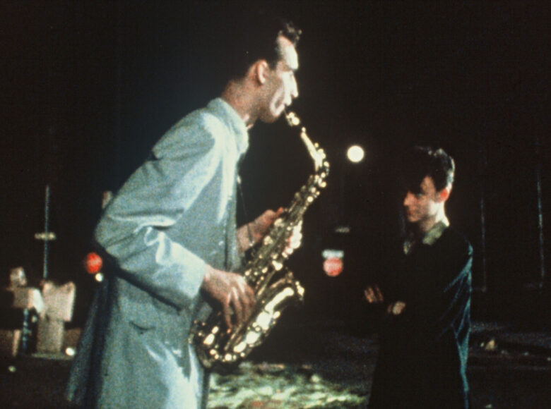 Na zdjęciu dwóch mężczyzn. Jeden gra na saksofonie, drugi przygląda mu się z rękami skrzyżowanymi na piersiach.