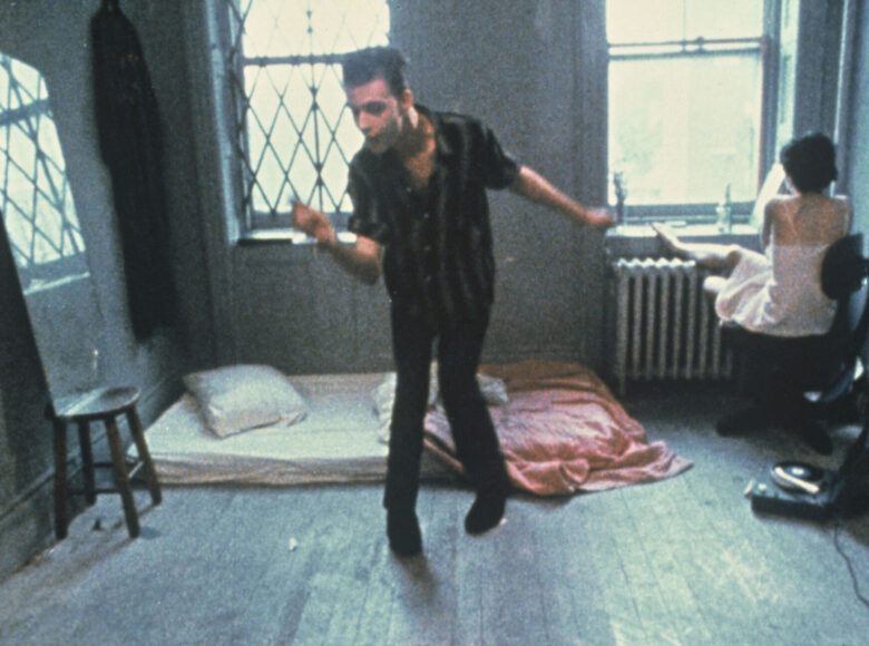 Na zdjęciu mężczyzna i kobieta w pokoju. Mężczyzna wydaje się tańczy na środku, po prawej stoi oparte o ścianę lustro, na podłodze leży materac. Kobieta siedzi tyłem do kadru, przed oknem.