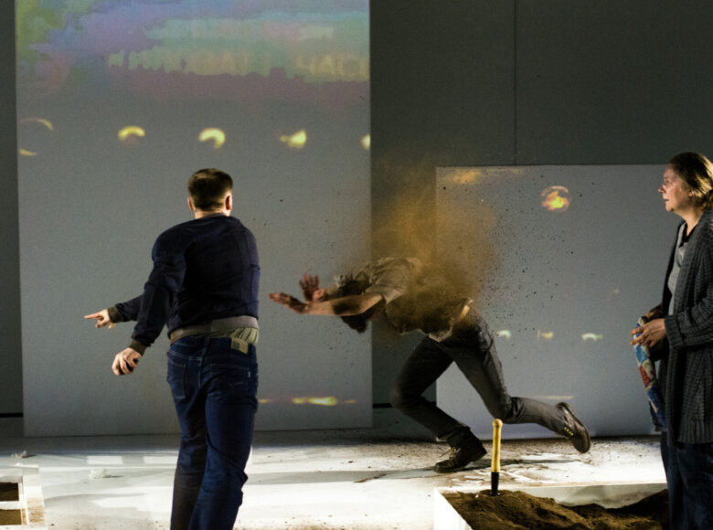 Dynamiczna scena. Na scenie stoi trójka ludzi. Kobieta stoi po prawej stronie i przygląda się szamotaninie dwójki mężczyzn. Jeden z nich jest odwrócony plecami do widowni i rzuca piaskiem w drugiego mężczyznę. Mężczyzna obrzucany piaskiem osłania twarz, potyka się.