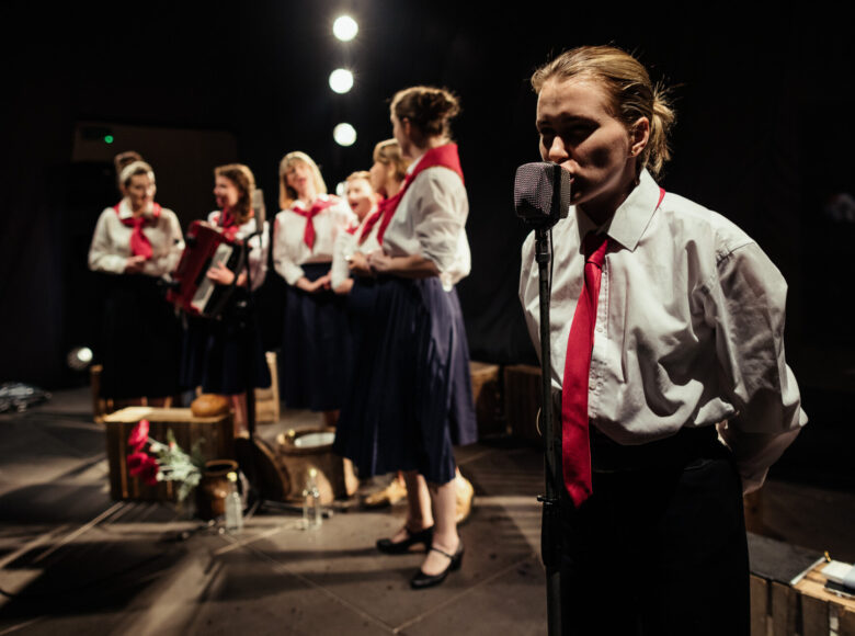 Zdjęcie. Scena ze spektaklu. Kobieta w białej koszuli i w czerwonym krawacie znajduje się na scenie i wygłasza mowę do mikrofonu.