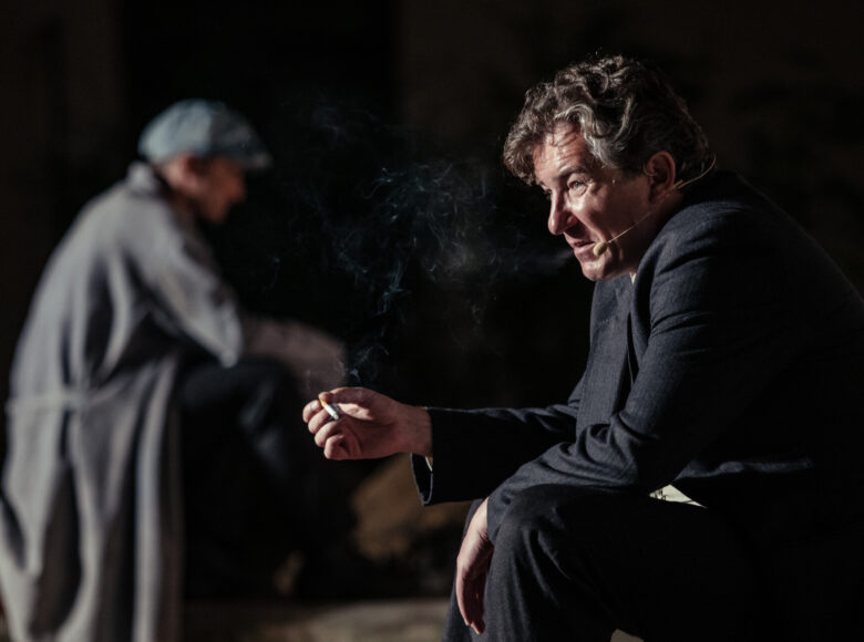 Zdjęcie. Scena ze spektaklu. Mężczyzna w ciemnej marynarce siedzi na skrzynce i pali papierosa.