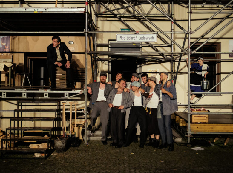 Zdjęcie. Scena ze spektaklu. Scenografia przedstawia plac budowy i rusztowania. Na scenie znajduje się roześmiana grupa budowlańców.