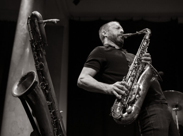 Zdjęcie w orientacji poziomej, czarno-białe. Na ciemnym tle, na pierwszym planie, z lewej strony widoczny jest saksofon, na statywie, zwrócony w lewą stronę. Z prawej strony znajduje się postać białego mężczyzny, w krótkich jasnych włosach. Mężczyzna jest widoczny w ujęciu amerykańskim, z profilu. W ręku trzyma saksofon. Kadr prezentuje mężczyznę podczas gry na instrumencie, jego ciało jest przechylone do tyłu. Za postacią mężczyzny widoczny jest talerz perkusyjny na statywie.