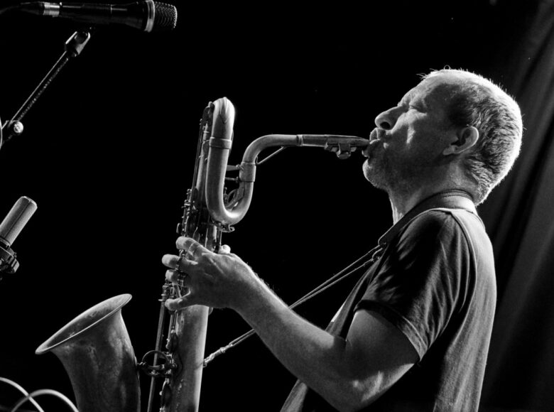Zdjęcie w orientacji poziomej, czarno-białe. W prawej części fotografii znajduje się postać białego mężczyzny o blond krótkich włosach i kilkudniowym zaroście. Mężczyzna jest widoczny z profilu, w ujęciu do pasa. W rękach trzyma saksofon zawieszony na pasku, na szyi. Ustnik instrumentu trzyma przy otwartych ustach. W górnym lewym rogu znajduje się mikrofon na statywie, zwrócony w kierunku mężczyzny, poniżej widać drugi mikrofon. Na zdjęciu dominuje ciemne tło.
