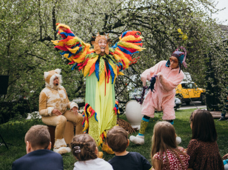 Zdjęcie przedstawia scenę ze spektaklu plenerowego "Fju Fju". Troje aktorów jest przebranych za ptaki. Na trawie siedzą dzieci i oglądają przedstawienie.