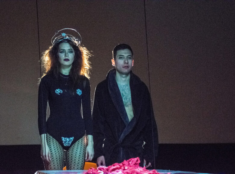 Kobieta z burzą loków stoi na baczność w towarzystwie niższego mężczyzny w szlafroku. Oboje są ubrani na czarno. Na ich twarzach nie widać emocji. Przed nim stół, na którym leżą wnętrzności ludzkie.