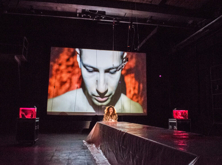 Kobieta siedzi samotnie u szczytu stołu. Za nią wyświetla się jasna twarz mężczyzny, który spogląda w dół.