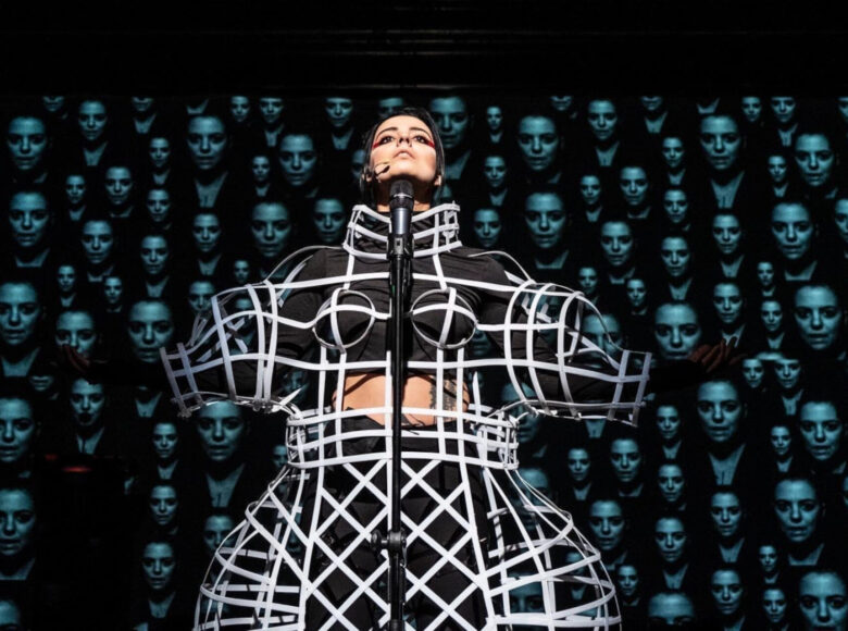 Kobieta w futurystycznym stroju stoi przed mikrofonem z uniesionymi ramionami. Jej twarz jest skierowana ku górze. Sylwetka kobiety jest mocno zniekształcona przed ubiór. W tle wyświetlane są setki identycznych twarzy.
