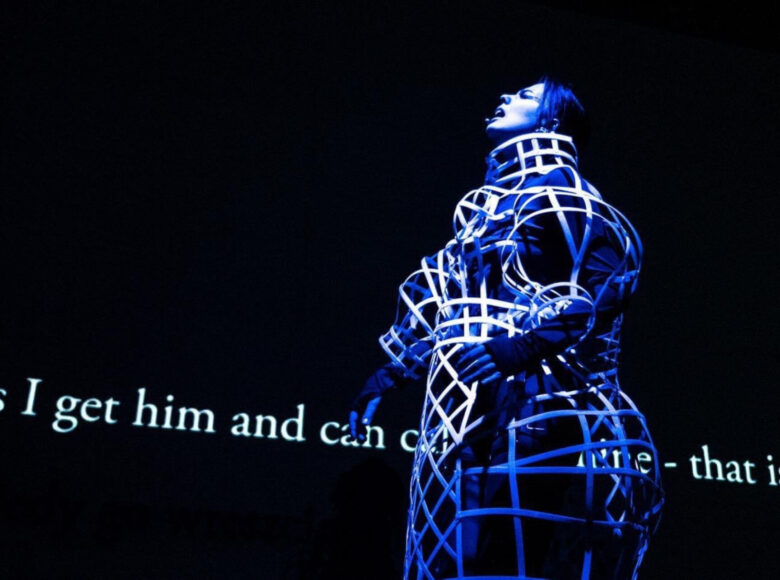 Postać kobiety oświetlona na niebiesko. Jej twarz jest zwrócona ku górze, jej wargi są rozchylone. W tle czarny ekran i wyświetlane białe napisy w języku angielskim.