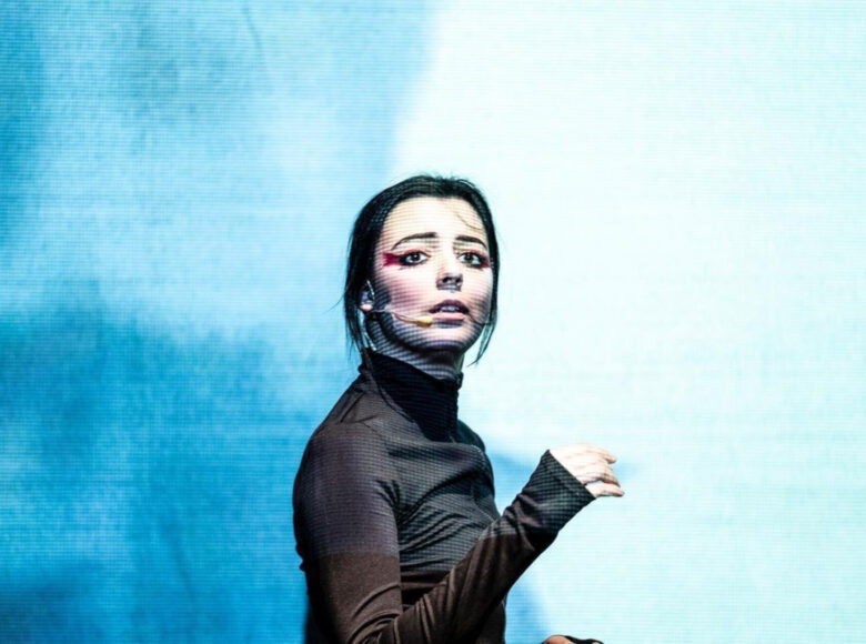 Młoda kobieta z ciemnymi włosami w czarnym golfie. Jej ciało jest obrócone w prawą stronę, a twarz skierowana do kamery. Ma lekko rozchylone wargi. Szaro-niebieskie światło wyświetlane przez projektor pada na sylwetkę kobiety.