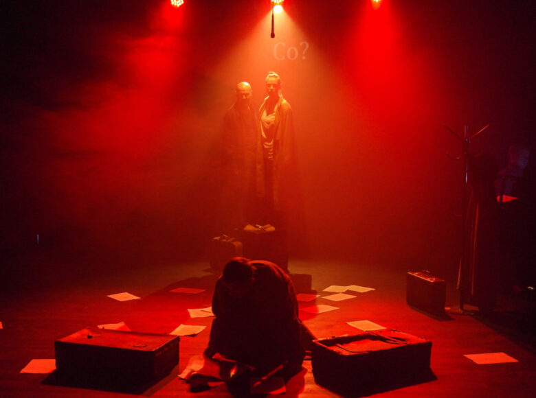 Zdjęcie. Na scenę pada czerwone światło, nad podłoga unosi się dym. Mężczyzna klęczy na ziemi i próbuje pozbierać rozrzucone kartki. Za nim, na podwyższeniu stoją dwie osoby, które na niego patrzą.