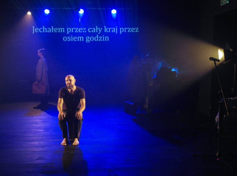 Zdjęcie. Na scenę pada niebieskie światło. Mężczyzna z ciemnym ubraniu siedzi na stołku. Jest bosy, ręce trzyma na kolanach. Za nim wyświetla się napis: Jechałem przez cały kraj przez osiem godzin.