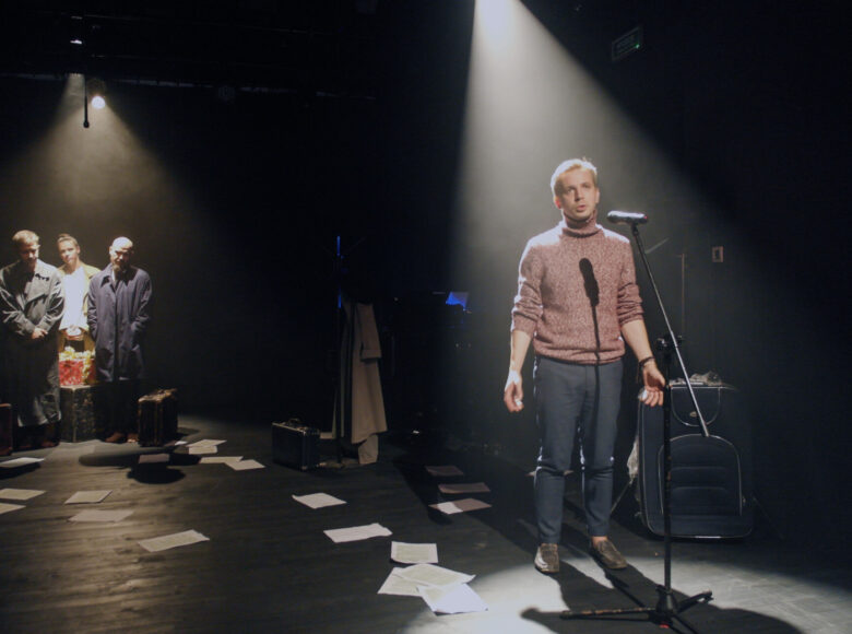 Zdjęcie. Na podłodze rozrzucone są białe kartki. Na scenie znajduje się grupa ludzi. Młody mężczyzna stoi przy mikrofonie i śpiewa. Pada na niego snop światła.