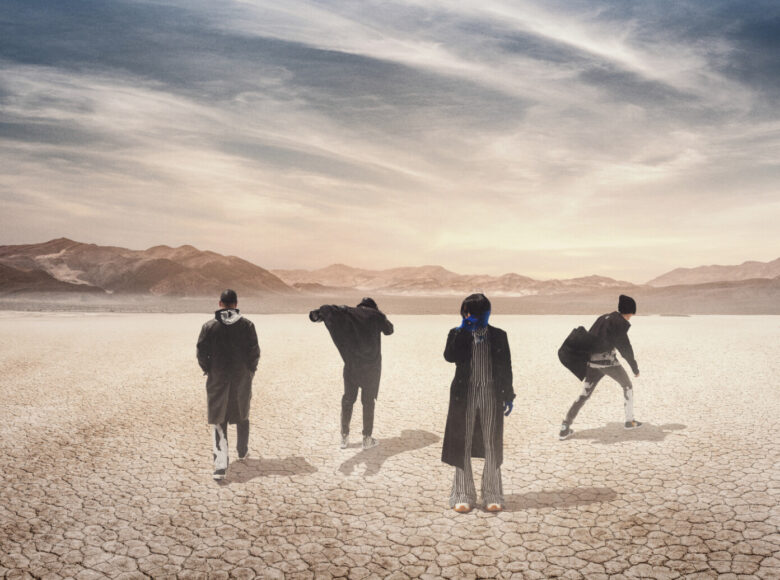 Zdjęcie. Grupa ludzi w szarych kostiumach i czarnych płaszczach znajduje się na pustyni. W oddali znajdują się skaliste wzniesienia.