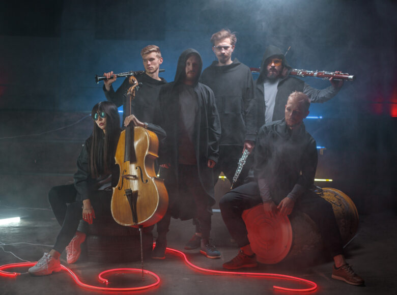 Fotografia przedstawia sześcioro osób w ciemnym, zadymionym pomieszczeniu, z przewagą niebieskiego i szarego koloru. Postaci widoczne są w otoczeniu instrumentów, co wskazuje na to, że tworzą zespół muzyczny. Niektórzy z nich trzymają je w swoich dłoniach. Cztery osoby uwiecznione zostały w pozycji stojącej, a inne dwie w pozycji siedzącej. Wszyscy mają na sobie czarne, bądź szare ubrania. Na podłodze znajduje się kontrastujący z nimi czerwony, świecący kabel/wąż. Dwie spośród widocznych osób posiadają okulary przeciwsłoneczne.