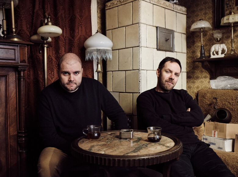 Zdjęcie. Dwóch mężczyzn siedzi przy małym stoliku, na którym lezą filiżanki z kawą. Pokój, w którym znajdują się mężczyźni jest urządzony w stylu vintage. Lampy mają białe abażury z frędzlami. okna zasłonięte są brązowymi kotarami. Na półce stoi biała figurka, porcelanowego słonia.