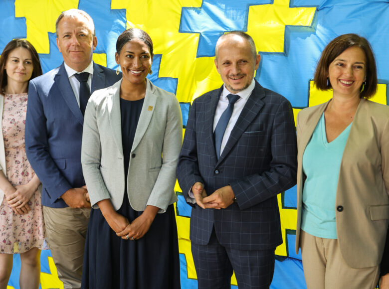 Zdjęcie z konferencji prasowej. Grupa ludzi znajduje się w parku. Za nimi znajduje się totem z niebiesko-żółtego materiału.