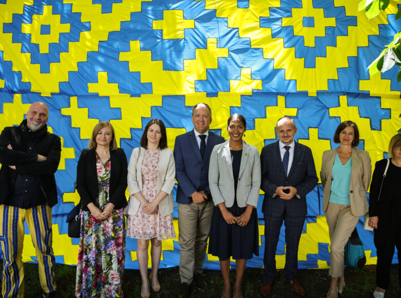 Zdjęcie z konferencji prasowej. Grupa ludzi znajduje się w parku. Za nimi znajduje się totem z niebiesko-żółtego materiału.