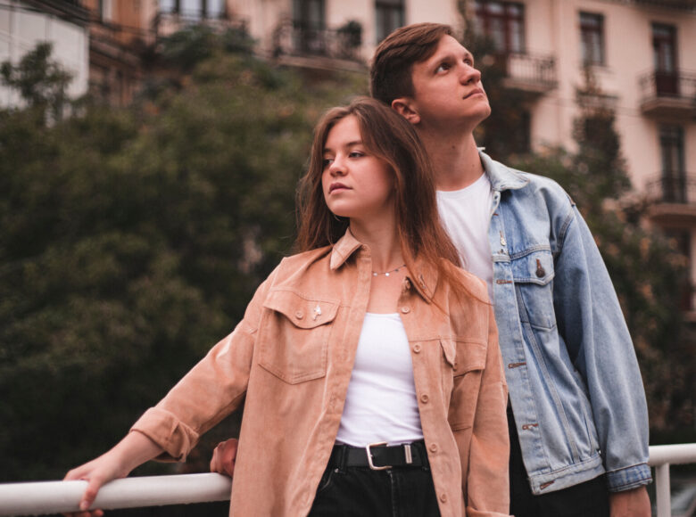 Zdjęcie. Młody mężczyzna i kobieta stoją razem przy barierce. Za nimi znajdują się bloki mieszkalne i drzewa.