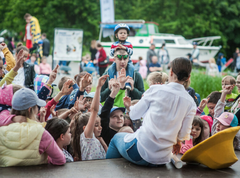 Zdjęcie. Grupa dzieci stoi pod sceną i unosi ręce go góry. Za nimi znajduje się zielony park i łódka na lawecie.