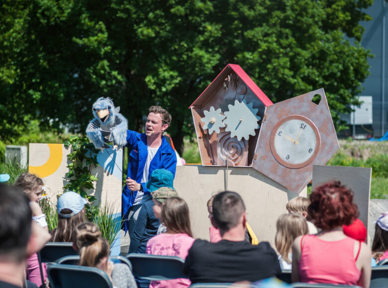 Zdjęcie. W parku rozstawiona jest scena, na której znajduje się scenografia do spektaklu, przedstawiająca zegar z kukułką. Aktor w niebieskim płaszczu występuje z kukiełką przed grupą dzieci.