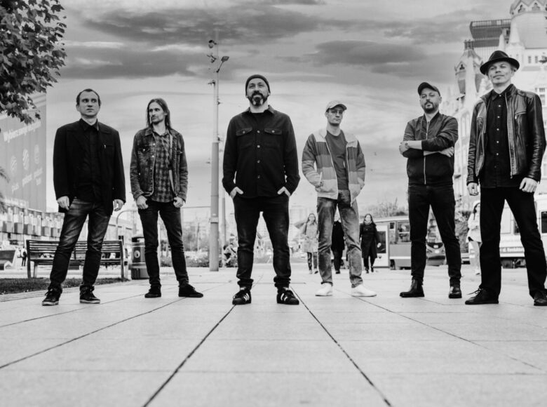 Fotografia przedstawia sześciu mężczyzn na tle fragmentu miasta z przechodniami. Zdjęcie widoczne jest w tonacji czano-białej. Cztery postaci mają nakrycie głowy w formie czapki, czapki z daszkiem lub kapelusza. Na ich ubraniach przeważa czerń. Tylko trzech z uwiecznionych mężczyzn patrzy prosto w obiektyw, a wzrok reszty postaci jest utrwalony w innych kierunkach. Mężczyzna z najbujniejszą brodą jest umieszczony na pierwszym planie fotografii.