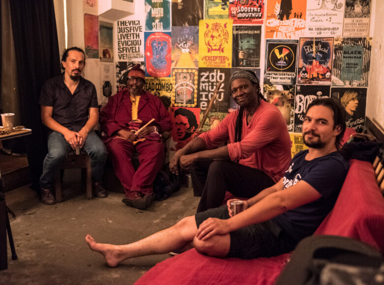 Fotografia przedstawia czterech mężczyzn widocznych na tle kolorowej ściany, zawierającej wizualne materiały przypominające plakaty. Wszystkie uwiecznione na zdjęciu postaci są w pozycji siedzącej. W pomieszczeniu z przewagą białych ścian panuje nieporządek. Dwóch mężczyzn ma na sobie ubrania w stonowanych kolorach – czarne, szare i granatowe, a pozostałych dwóch wyróżnia się strojem przybierającym intensywne barwy – czerwień oraz róż.