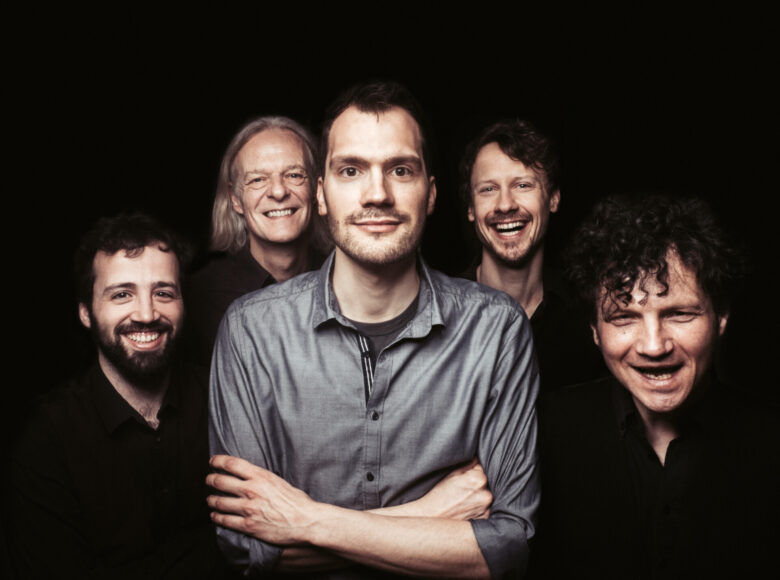 Fotografia przedstawia pięciu mężczyzn na czarnym tle. Jeden z nich jest widoczny na pierwszym planie z założonymi rękami i jako jedyny nie ma czarnego stroju. Poza tym, na twarzach otaczających go postaci dostrzec można szerokie uśmiechy.