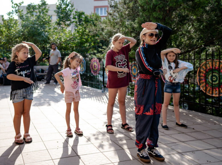 Zdjęcie. Grupa dzieci stoi w rzędzie i tańczy, naśladując ruchy osoby, która stoi przed nimi.