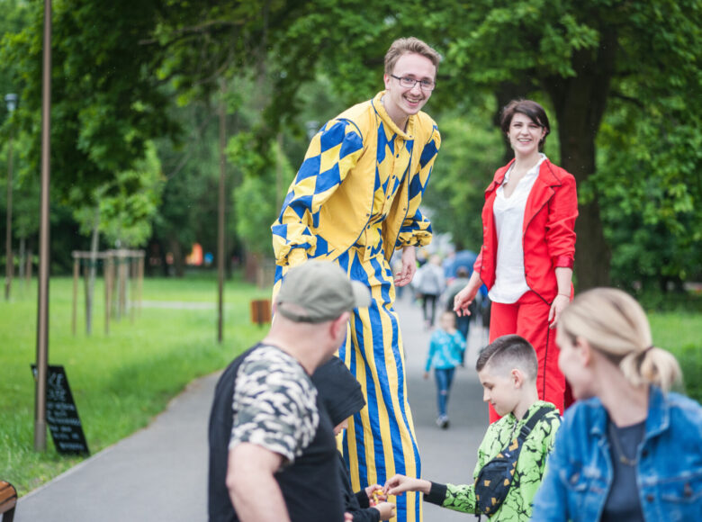 Zdjęcie. Ludzie znajdują się w parku i przyglądają się akrobatom, którzy chodzą na szczudłach.