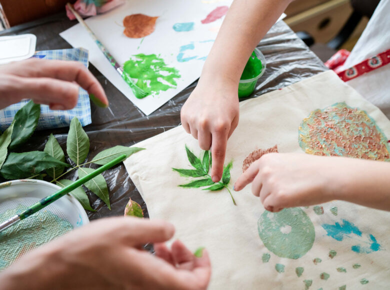 Zdjęcie. Na stole lezą kartony i farby. Dzieci malują liście farbami odbijają je na na kartonach.