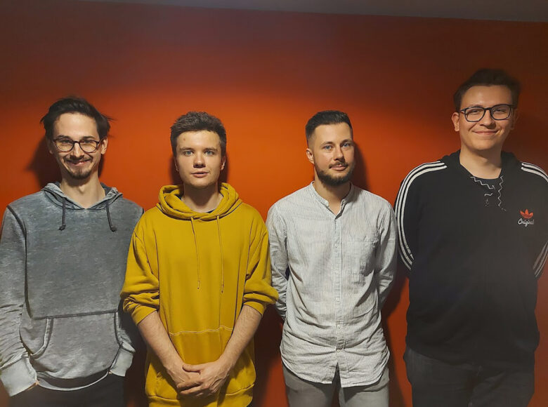 Zdjęcie. Przedstawia czterech młodych mężczyzn. Stoją pod pomarańczowa ściana i pozują do zdjęcia. Są uśmiechnięci.