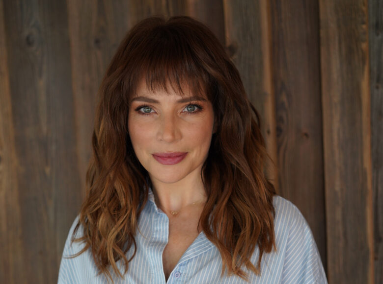 Zdjęcie. portret Anny Dereszowskiej. Kobieta stoi na tle drewnianej ściany. Uśmiecha się.