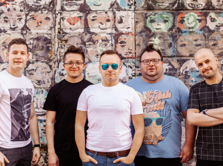 Zdjęcie. Ściana pokryta jest graffiti przedstawiającym powtarzającą się tą samą twarz. Przed ścianą stoi 5 mężczyzn. To członkowie zespołu Klezmafour.