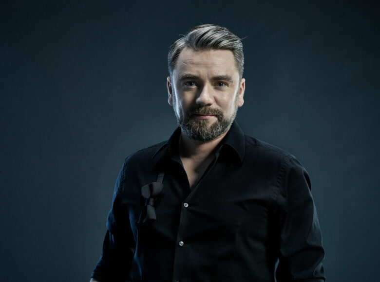 Zdjęcie. Portret wokalisty Kuby Badacha. Mężczyzna jest ubrany w czarna koszulę. Ma pogodny wyraz twarzy.