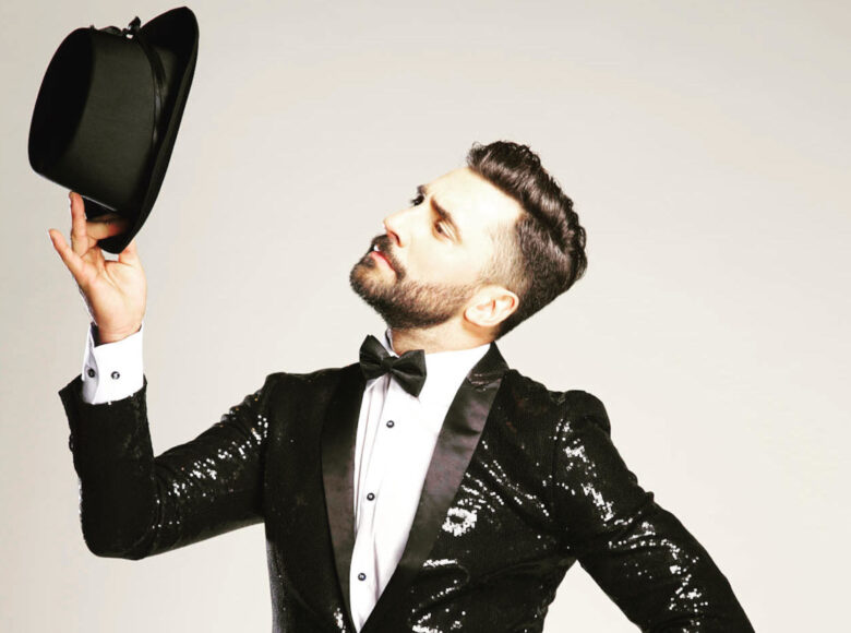 Zdjęcie. Przedstawia mężczyznę w ciemnej marynarce w cekiny. Mężczyzna ma na szyi zawiązaną muszkę. W ręce trzyma kapelusz.