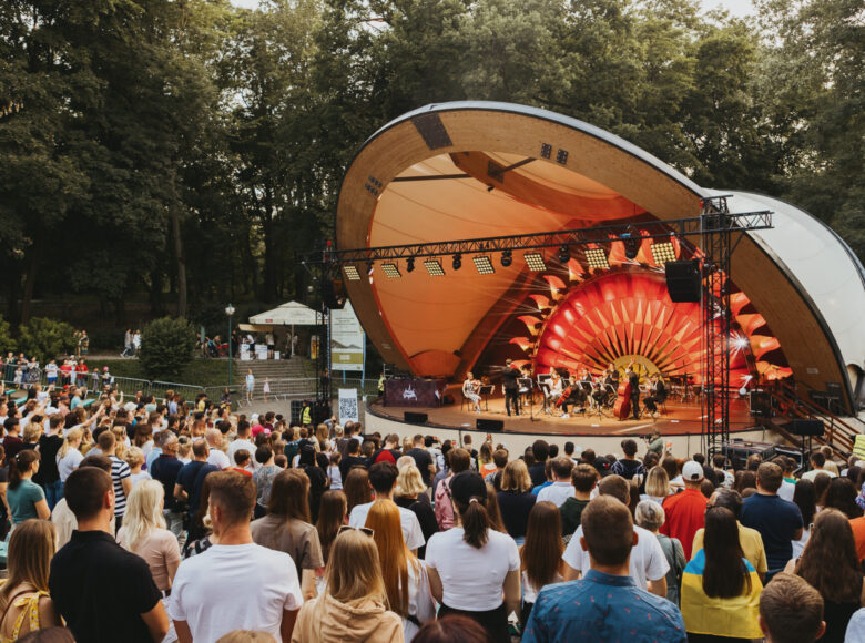 Szerokie ujęcie muszli koncertowej w ogrodzie saskim wraz z publicznością podczas koncertu orkiestry symfonicznej. Na scenie siedzą młodzi muzycy z instrumentami smyczkowymi, przed nimi stoi dyrygent. Scena jest oświetlona żółto-pomarańczowym światłem.
