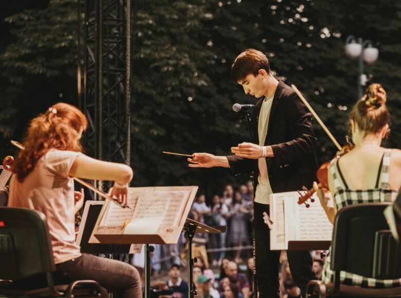 Zdjęcie przedstawia grupę muzyków na scenie Muszli Koncertowej w ogrodzie saskim. Dwie młode kobiety siedzą przed pulpitem z nutami, są zwrócone do nas plecami i grają na skrzypcach. Przed nimi stoi młody dyrygent.