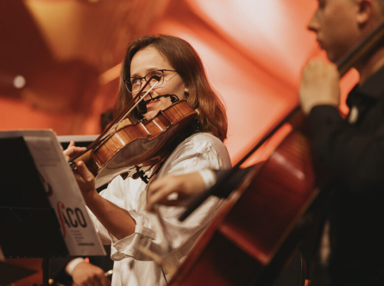 Zdjęcie przedstawia młodą kobietę ubraną w białą bluzkę, która siedzi na scenie i gra na skrzypcach podczas koncertu. Przed nią stoi pulpit z nutami. Po prawej stronie znajduje się nieostry zarys postaci męskiej grającej na wiolonczeli.