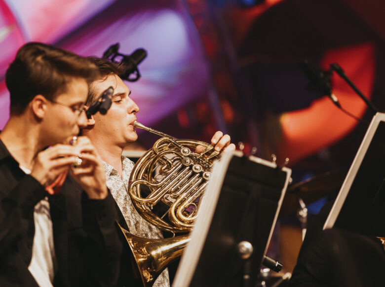 Zdjęcie przedstawia dwóch młodych mężczyzn na scenie podczas koncertu orkiestry symfonicznej. Jeden z nich gra na flecie poprzecznym, drugi na waltorni. Siedzą przed pulpitami z nutami.