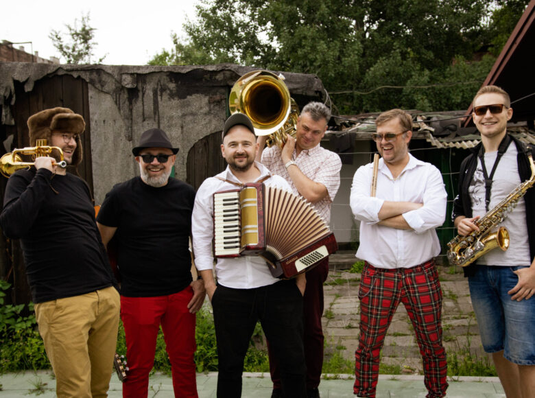 Zdjęcie. Przed drewnianymi budynkami stoi grupa muzyków ze swoimi instrumentami. To członkowie zespołu PropaBanda. Muzycy są uśmiechnięci.