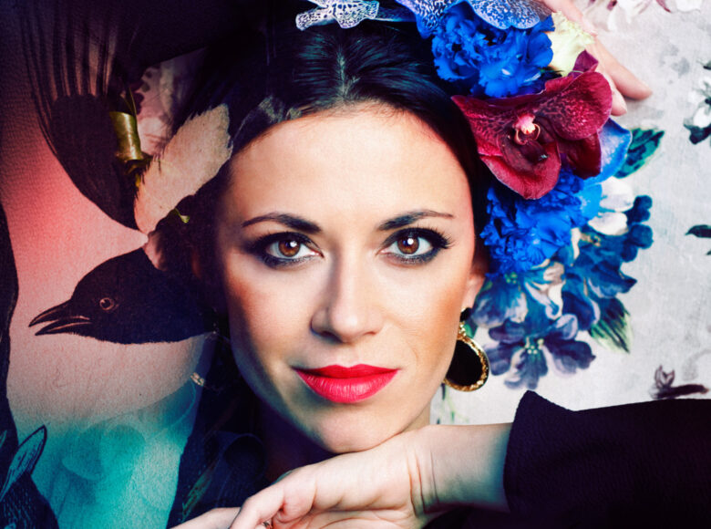 Zdjęcie. Portret kobiety. Ma pomalowane jaskrawą szminką usta. Kobieta ma wpięte we włosy niebieskie i czerwone orchidee.