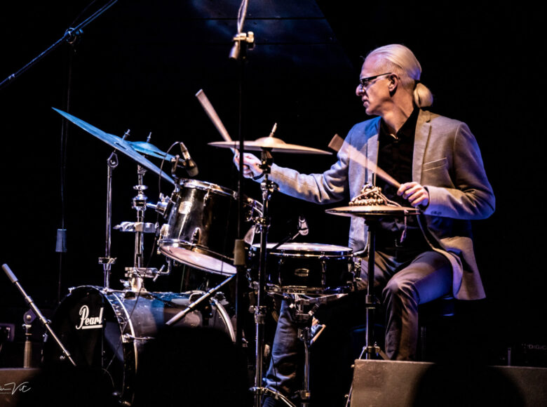 Zdjęcie. Przedstawia mężczyznę o siwych włosach, związanych na karku. Mężczyzna gra na perkusji.